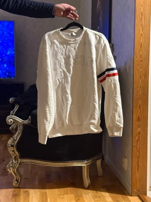 Vit sweatshirt från Moncler - Vit sweatshirt från Moncler med broderad logga på bröstet och coola ränder i blått, vitt och rött på ena ärmen. Rund hals och långa ärmar, tillverkad i mjuk bomull. Perfekt för dig som gillar stilrena och sportiga vibes.