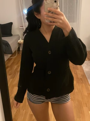 Svart cardigan från 157, XS - Den virala Svart cardigan från 157 i storlek XS. Koftan har v-ringning, stora knappar framtill och är ribbstickad vid ärmslut och midja. Knappt använd. Finns likadan i brun. Storlek xs på den med. 