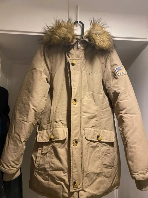 Beige parkajacka med päls från Svea - Snygg beige parkajacka från Svea med fuskpäls på huvan, broderad logga på bröstet och patch på ärmen. Jackan har dragkedja och knappar framtill, två stora fickor med lock och quiltad insida. Perfekt för kalla dagar och har en klassisk look.