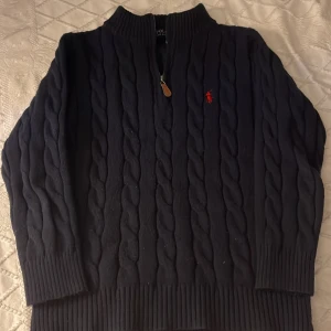 Svart stickad tröja Polo Ralph Lauren - Svart kabelstickad tröja från Polo Ralph Lauren med halv dragkedja och röd broderad logga på bröstet. Tröjan har ribbade muddar och hög krage, perfekt för kyliga dagar. Klassisk och stilren design som funkar till många outfits.