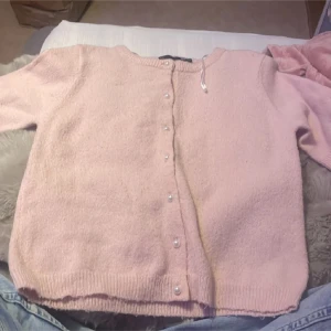 Ljusrosa stickad kofta från 157 - Mysig ljusrosa stickad kofta från 157 i storlek XS. Koftan har rund hals, långa ärmar och pärlknappar framtill. Ribbstickade muddar vid ärmslut och nederkant ger en snygg finish. Perfekt för lager-på-lager under kyliga dagar.