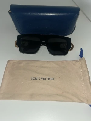 Svarta  Louis Vuitton   solglasögon med fodral - Säljer ett snyggt blått fodral och solglasögon från Louis Vuitton, perfekt för att förvara dina solglasögon. Fodralet är tillverkat i skinn och har en stilren design med präglad logga. Medföljer även en beige tygpåse med Louis Vuitton-tryck. 