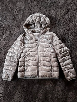 Grå pufferjacka från Uniqlo  - Säljer en grå pufferjacka från Uniqlo i storlek L men passar mer som Xs . Jackan har huva, dragkedja framtill och är quiltad med horisontella sömmar. Materialet är syntet och jackan är lätt och smidig att bära. Perfekt för kyliga dagar och enkel att matcha med olika outfits. Hör av er om ni har några frågor!☺️
