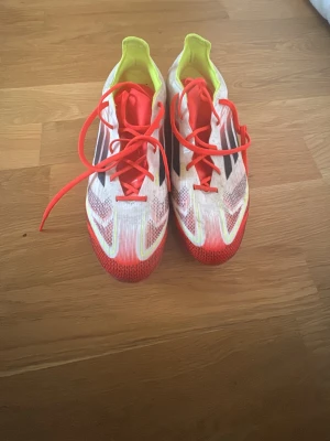 Adidas vita och röda fotbollsskor - Säljer ett par Adidas fotbollsskor med vita och röda detaljer, svarta ränder på sidan och neonfärgade inslag. Skorna har snörning och platt sula med dobbar för gräsplan. Ovandelen är i syntetmaterial och insidan har en neonfärgad finish. Perfekt för dig som älskar fotboll.