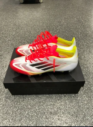 F50 elite  - Säljer ett par Adidas fotbollsskor med snygg design i vitt, rött och gult. Skorna har svarta ränder på sidan, röda snören och ventilerande meshmaterial. Perfekta för dig som vill sticka ut på planen med både stil och funktion.
