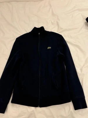 Mörkblå ziptröja från Lacoste Sport - Snygg mörkblå ziptröja från Lacoste Sport med klassisk ståkrage och dragkedja hela vägen. Tröjan har det ikoniska gröna krokodilmärket på bröstet och två sidofickor. Perfekt för dig som gillar sportig och clean stil.