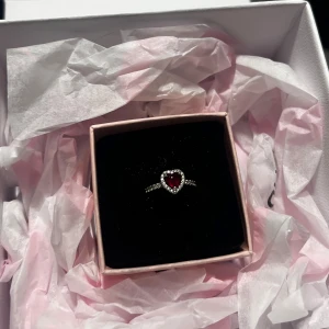 Ring med hjärtformad röd sten - Elegant ring från Pandora med en hjärtformad röd sten omgiven av gnistrande stenar. Perfekt för att ge en romantisk touch till din stil.  presentasken ingår. Jag köpte den för ett tag sedan men, den har bara kommit till användning 2 gånger pga att jag bytte till guld. Original priset är 900 men jag säljer den för 400, dock så kan man buda.