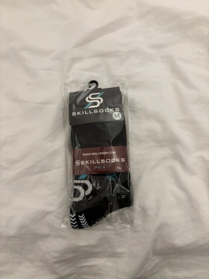 Svarta sportstrumpor från SkillSocks - Svarta sportstrumpor från SkillSocks Pro:2 med vita och blå detaljer samt grepp på undersidan. Tillverkade i en blandning av merinoull, nylon och elastan för bästa komfort och andningsförmåga. Perfekta för träning och sportiga aktiviteter.