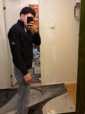 Svart stickad kofta från Stone Island - Svart ribbstickad kofta från Stone Island med dragkedja framtill och klassisk logopatch på vänster ärm. Koftan har hög krage och långärmad passform, perfekt för dig som gillar stilrena och tidlösa plagg. Materialet är mjukt och skönt stickat.