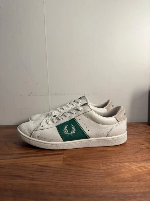 Fred Perry vita sneakers  - Säljer ett par vita Fred Perry sneakers med grönt parti på sidan och broderad logga. Skorna har snörning, rund tå och platt sula. Materialet är skinn med detaljer i mocka på hälen där Fred Perry är broderat i grönt. Perfekta för dig som gillar klassisk stil med en sportig touch.