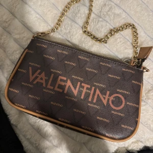 Brun handväska från Valentino - Säljer en snygg brun handväska från Valentino med guldigt kedjehandtag och dragkedja. Väskan har ett mönster med Valentino-loggor över hela ytan och kontrasterande beige kant. Perfekt för dig som gillar stilrena accessoarer med lyxig känsla.
