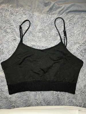 Svart croppad linnetopp med smala band - Säljer en svart croppad linnetopp med smala, justerbara axelband. Toppen har en enkel och stilren design med ribbad kant nedtill. Materialet känns mjukt och stretchigt, perfekt för att matcha med jeans eller kjol. Passformen är tight och toppen är ärmlös.