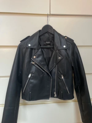 Svart skinnjacka från Pull&Bear - Snygg svart skinnjacka från Pull&Bear. Jackan har silvriga dragkedjor, bred krage och axelklaffar. Storleken är M men jag är XS och den passar mig perfekt. Skulle tro att den passar dig som har XS/S💕