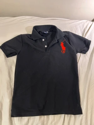 Svart polo tröja Ralph Lauren - Svart polotopp från Polo Ralph Lauren i 100% bomull. Klassisk krage, kortärmad och med tre vita knappar framtill. Stor röd broderad logga på bröstet och röd siffra på ärmen. Perfekt för en clean och sportig look.