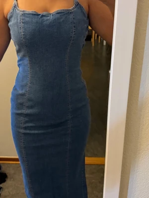 Blå jeansklänning från Bershka - Säljer en blå lång jeansklänning från Bershka med smala axelband och figurnära passform. Klänningen har en snygg slits på sidan och markerade sömmar som ger en cool look. Perfekt för dig som gillar denim och vill ha något trendigt i garderoben.