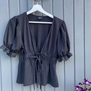 Svart blus med knytning - Supersnygg svart blus från Gina Tricot med djup v-ringning, puffiga korta ärmar och knyt i midjan. Blusen har en lätt struktur i tyget och volang vid ärmslut och midja. Perfekt för dig som gillar trendiga detaljer och markerad midja.