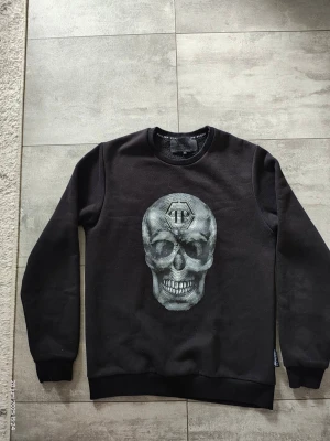 Svart sweatshirt med döskalle från Philipp Plein - Cool svart sweatshirt från Philipp Plein med ett stort silverfärgat döskalletryck på framsidan och broderad logga med årtal på ryggen. Tröjan har rund hals, ribbade muddar och är tillverkad i mjuk bomull. Perfekt för dig som gillar edgy streetstyle.