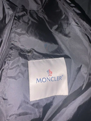 Svart vindjacka från Moncler - Svart vindjacka från Moncler med huva och dragkedja. Jackan har snygga röda och vita ränder på huvan och Moncler-logga på bröstet. Materialet är glansig polyester och jackan har snörning vid huvan för extra stilpoäng.
