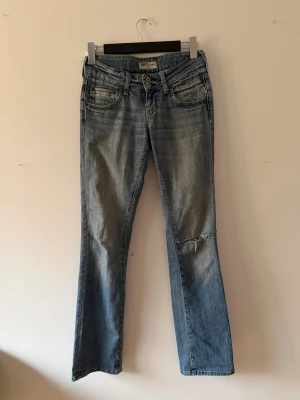 Blå bootcut jeans från Lee - Säljer ett par ljusblå bootcut jeans från Lee med slitningar och en trasig detalj vid knät. Jeansen har klassisk femficksdesign, dubbla knappar i midjan och är tillverkade i jeansmaterial. Perfekt för dig som gillar en avslappnad och cool stil.
