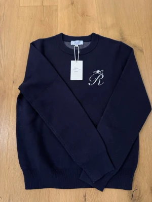 Mörkblå stickad tröja från Rêveur Atelier - Snygg mörkblå stickad tröja från Rêveur Atelier med vit broderad logga på bröstet och stor text på ryggen. Rund hals och långa ärmar, perfekt för dig som gillar stilrena detaljer och fransk vibe. Ribbstickade muddar vid ärmslut och nederkant. Mått: över bröstet 50 cm, ärm yttermått 63 cm, ärm innermått 48 cm, från hals till tröjans nederkant 63 cm, Storlek XS. Köpt för 1013 kr, oanvänd med märkeskappen kvar, kvitto finnes. 