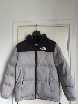 Grå och svart pufferjacka från The North Face - Säljer en grå och svart pufferjacka från The North Face med hög krage och dragkedja framtill. Jackan har två sidofickor med dragkedja och logga på bröstet. Perfekt för kalla dagar och i storleken Medium. Finns att hämta upp i centrala Stockholm.