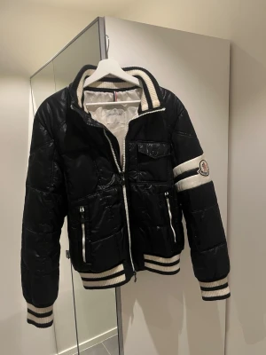 Svart pufferjacka från Moncler - Tja, säljer en svart pufferjacka från Moncler med vita detaljer och ribbade muddar. Jackan har dragkedja framtill, fickor med dragkedja och en vit Moncler logga på ärmen.