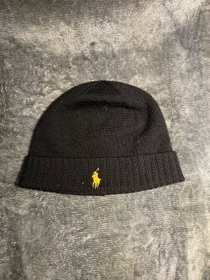 Svart mössa från Polo Ralph Lauren - Svart ribbstickad mössa från Polo Ralph Lauren i mjuk och tvättbar merinoull. Klassisk design med gul broderad logga framtill. Perfekt för kalla dagar och enkel att matcha med din stil. Pris går att diskutera 