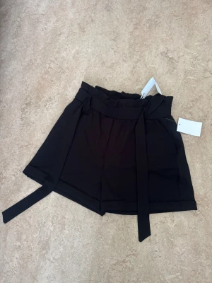 Svarta högmidjade shorts med bälte - Säljer ett par svarta shorts med hög midja och breda ben. De har ett snyggt knytbälte i midjan och diskreta fickor bak. Materialet känns mjukt och har en lätt struktur, perfekt för en chill och stilren look.