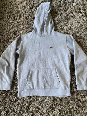 Grå hoodie från Lacoste med huva - Säljer en stilren grå hoodie från Lacoste med klassisk krokodillogga på bröstet. Tröjan har huva, magficka och ribbade muddar vid ärmslut och nederkant. Perfekt för chill dagar och enkel att matcha med jeans eller joggers.
