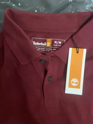 Vinröd långärmad Timberland skjorta - Säljer en vinröd långärmad skjorta från Timberland i slim fit. Skjortan har klassisk krage, två knappar framtill och broderad Timberland-logga på bröstet. Materialet är 100% bomull och känns mjukt mot huden. Perfekt för dig som gillar stilrena och enkla plagg.