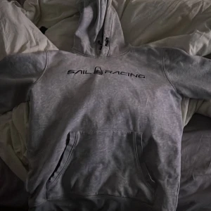 Grå hoodie från Sail Racing - Säljer en grå hoodie från Sail Racing med svart logga på bröstet. Tröjan har huva med dragsko och en stor magficka framtill. Insidan är mjuk och fleecefodrad, perfekt för kyliga dagar. Klassisk och stilren design som funkar till det mesta.