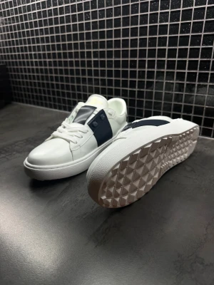 Valentino Opens - Snygga Valentino Open sneakers i vitt skinn med bred marinblå läderdetalj över mitten. Klassisk rund tå, platt sula och vita snören. Guldtext på plösen och diskreta nitar på sidan ger en lyxig känsla. Perfekta för dig som vill ha en clean men exklusiv look.