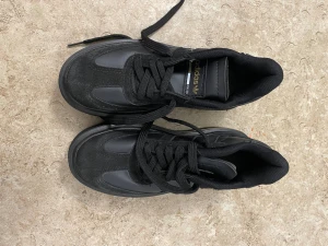 Svarta Adidas sneakers med plattform - Säljer ett par svarta Adidas sneakers med platt sula och diskret gulddetalj runt sulan. Skorna har klassiska tre ränder på sidan, snörning och är tillverkade i en mix av mocka och syntetmaterial. Adidas-logga i guld på hälen och tungan ger en snygg touch.