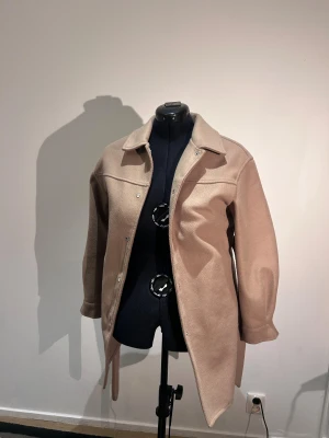 Beige kappa i ullmix - Snygg beige kappa med klassisk krage och tryckknappar framtill. Jackan har en rak och loose passform, perfekt att slänga över en hoodie eller t-shirt. Materialet är mjukt och har en ullkänsla som ger extra värme under kyliga dagar.