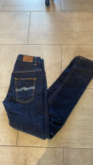Nudie Jeans Thin Finn blå jeans - Säljer ett par mörkblå Nudie Jeans Thin Finn med klassisk femficksdesign och snygga kontrastsömmar. Jeansen är tillverkade av 98% ekologisk bomull och 2% elastan för extra komfort. Smal passform och stilren look med broderad bakficka. 30/32