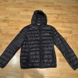 Svart pufferjacka EA7 Emporio Armani - Svart pufferjacka från EA7 Emporio Armani med guldfärgat logotryck på bröstet. Jackan har huva, dragkedja framtill och quiltad design. Perfekt för dig som vill ha en snygg och lätt jacka med streetkänsla. Materialet är syntet och jackan är långärmad.