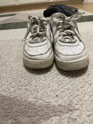 Nike Air Force 1 vita sneakers - Säljer ett par klassiska Nike Air Force 1 sneakers i vitt skinn med rund tå och platt sula. Skorna har snörning och perforerade detaljer på tån för extra ventilation. Swoosh-loggan syns tydligt på sidan och de har en låg siluett som passar till många stilar.