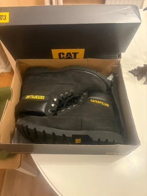 Svarta boots från Caterpillar - Säljer ett par svarta boots från Caterpillar med grov sula och snörning. Skorna har robust design, gul logga på sidan och plös, samt detaljer i skinn och mocka. Perfekta för dig som gillar chunky stil och vill ha något slitstarkt till hösten och vintern.