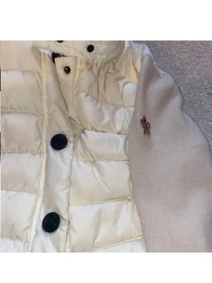 Vit Moncler Grenoble dunjacka - Säljer en snygg vit cardigan från Moncler med stickade beige ärmar och svarta knappar framtill. Jackan har puffad design och Moncler-logga broderad på ärmen. Perfekt för dig som vill ha en stilren och varm jacka med exklusiv känsla.