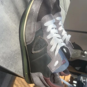 Philippe Model grå sneakers mocka - Snygga sneakers från Philippe Model i grå och mörkgrå toner med detaljer i vinrött. Skorna har en mix av mocka och skinn, vit snörning och en rund tå. Sidopanelen pryds av en broderad sköld. Perfekt för dig som gillar stilrena och coola sneakers.