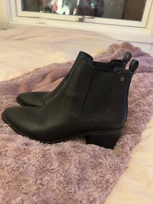 Svarta chelsea boots med klack - Säljer ett par svarta chelsea boots i skinn med spetsig tå och låg klack. De har elastiska paneler på sidorna och dragflikar bak för enkel påtagning. Perfekta för dig som gillar stilrena och klassiska boots med lite edge.