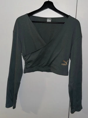 Grå omlottopp från Puma - Säljer en grå croppad omlottopp från Puma med långärmad design och ribbad struktur. Toppen har en snygg v-ringning och Puma-logga i guld på framsidan. Perfekt för dig som gillar sportiga och trendiga plagg.
