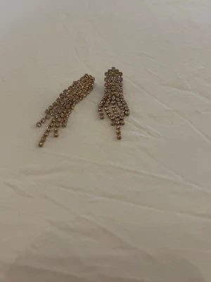 Örhängen  - Bilderna visar ett par glittriga örhängen i guldton med hängande strassdetaljer, men ingen väst finns med på bilderna. Använda 1 gång.