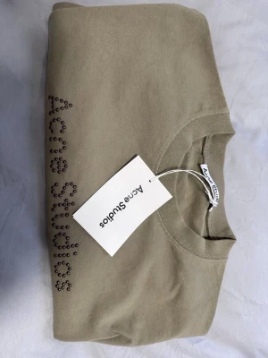 Beige t-shirt Acne Studios med pärlor - Snygg beige t-shirt från Acne Studios i 100% bomull. Framsidan har Acne Studios-loggan i pärldetaljer som ger en unik vibe. Klassisk rund hals och korta ärmar. Perfekt för dig som gillar stilrena plagg med en twist.