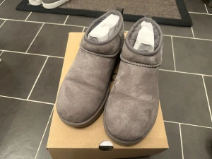 Ugg Classic Ultra Mini, Grå, EUR 41 - Säljer ett par grå UGG boots med låg profil och rund tå. Skorna är tillverkade i mjuk mocka och har en platt sula. UGG-loggan syns baktill och insidan är fodrad för extra komfort. Perfekt för dig som gillar chill och stilren look.