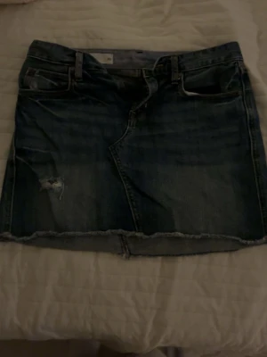 Blå jeanskjol från Gap 1969 XS - Säljer en blå jeanskjol från Gap 1969 i storlek XS. Kjolen har klassisk femficksdesign, rå kant nedtill och slitning på framsidan för en cool look. Passar dig som gillar denim och vill ha en kort kjol.