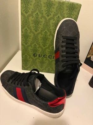 Gucci sneakers med röd och blå detalj - Säljer ett par svarta Gucci sneakers med klassiska röd och blå ränder på sidan. Skorna har snörning, vit platt sula och metallic-detaljer i rött och blått på hälen med Gucci-logga. Materialet är canvas med subtilt GG-mönster och skinn på hälen.