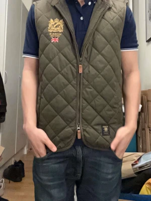 dunväst Morris XS - Säljer en olivgrön quiltad dunväst från Morris i storlek XS. Västen har snygga broderade detaljer med guldfärgat emblem och brittisk flagga på bröstet. Dragkedja framtill och klassisk sportig look. Perfekt för lager-på-lager under höst och vinter.