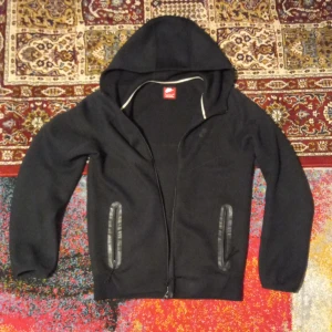 Svart Nike hoodie med dragkedja - Svart hoodie från Nike med huva och hel dragkedja framtill. Tröjan har två fickor med dragkedja och ett diskret Nike-logotryck på bröstet. Insidan har en mjuk känsla och huvan har en vit kant som kontrast. Perfekt för chill eller träning.