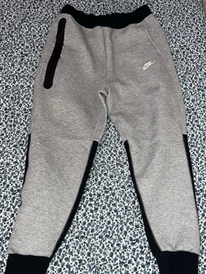 Nike Tech mjukisbyxor - Säljer ett par grå mjukisbyxor från Nike med svarta muddar och svarta paneler längs benen. Byxorna har en vit Nike-logga på framsidan och en svart dragkedja vid fickan. Sköna och stilrena, perfekta för chill eller träning. Helt ny aldrig använda. Storlek M. Pris kan diskuteras vid snabbaffär.